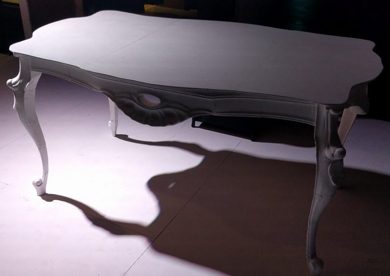 A printed table with scalloped edges and curved cabriole legs, lit from one side on a dark stage floor so it throws a long shadow.