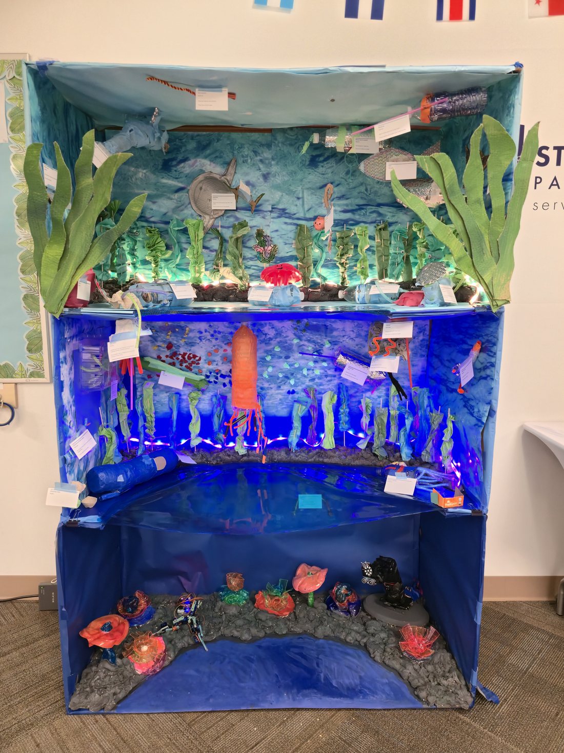 A three-level ocean diorama in a classroom. The sunlit top zone has kelp and fish made from recycled materials, the middle twilight zone glows blue with a large orange jellyfish and LED lighting, and the dark bottom zone holds fluorescent coral sculptures and small animatronic sea creatures on a grey sea floor. Handwritten labels are pinned throughout.