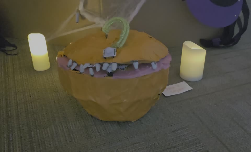 A papier-mache monster pumpkin with a motorized mouth, between two battery candles.