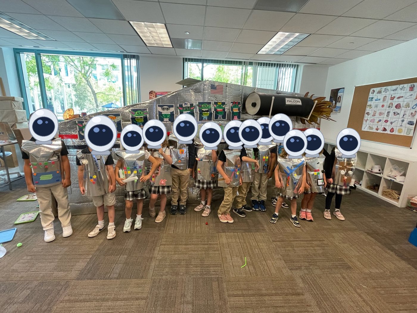 Thirteen kindergarteners in silver space suits stand in front of the U.S.S. Falcon, a foil-clad spaceship built in a classroom, with student alien artwork displayed on its hull. Cartoon space helmets are drawn over every child's head.